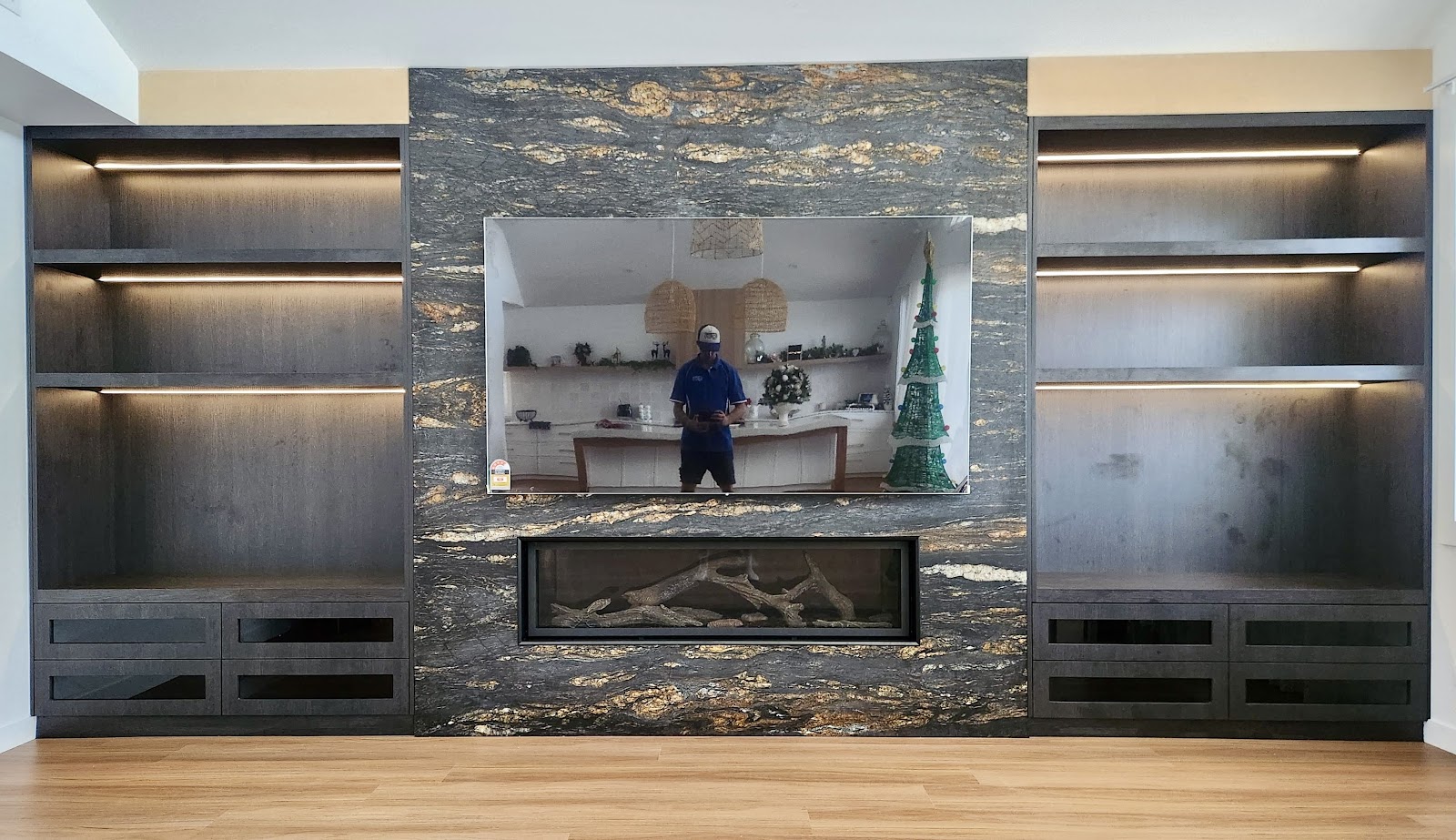 Dark joinery wall with lit open shelving either side of a stone fireplace
